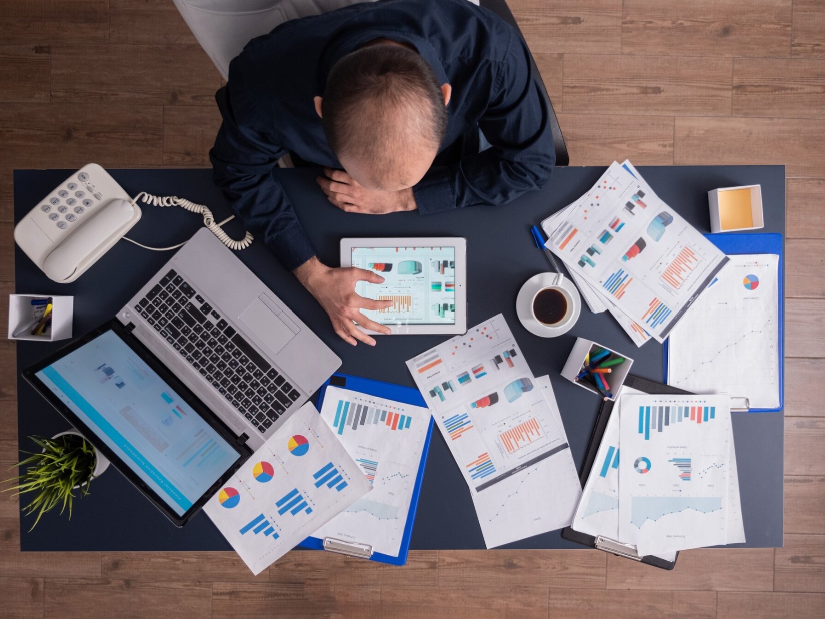 Homme consultant des tableaux de bord financiers et des graphiques de performance sur une tablette, entouré de rapports imprimés, d’un ordinateur portable et d’un café sur un bureau — scène illustrant l’analyse de données, la gestion de la performance ou la planification stratégique.