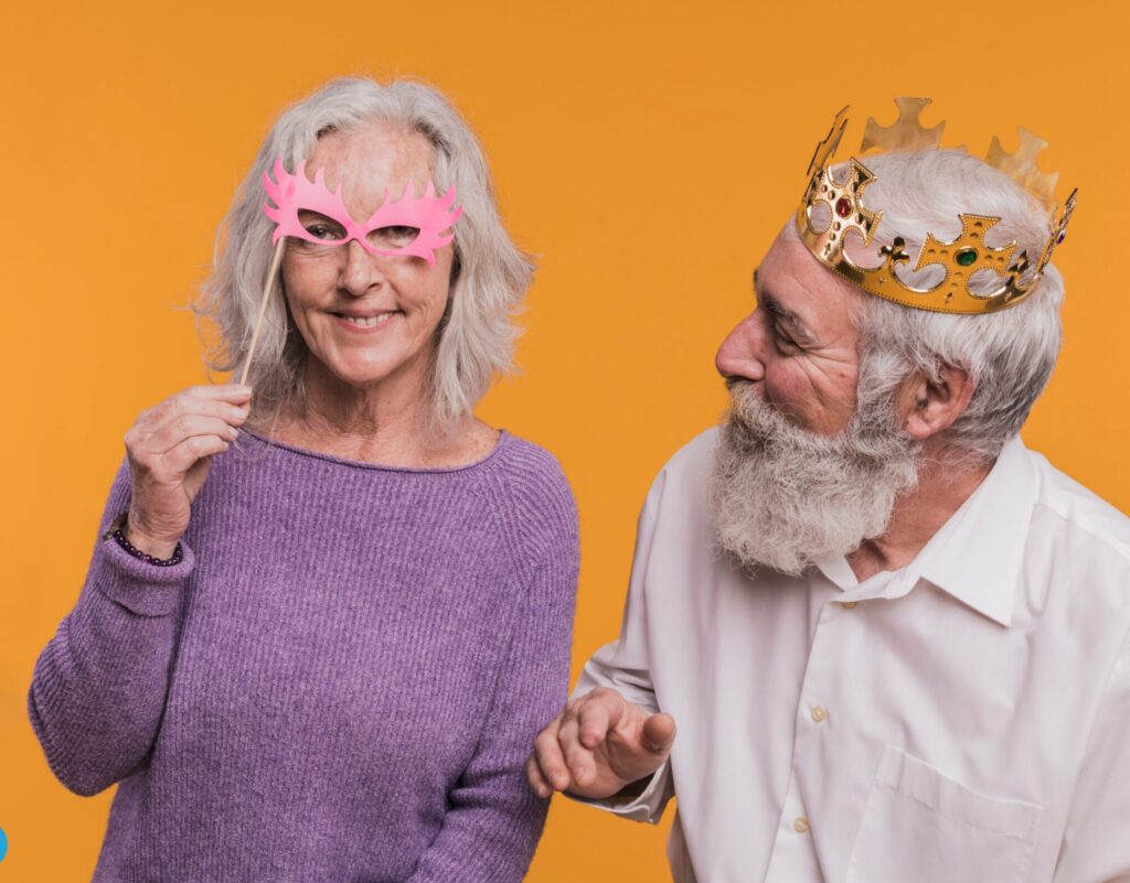 Portrait joyeux d’un couple senior sur fond orange vif. La femme sourit en tenant un masque rose festif, tandis que l’homme, couronné comme un roi, la regarde avec tendresse. Une scène ludique et complice, parfaite pour illustrer la vitalité et le bonheur après 60 ans.