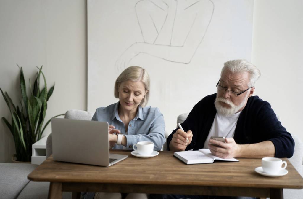 Deux adultes consultent un ordinateur et un carnet tout en échangeant des idées, assis à une table dans un intérieur lumineux.