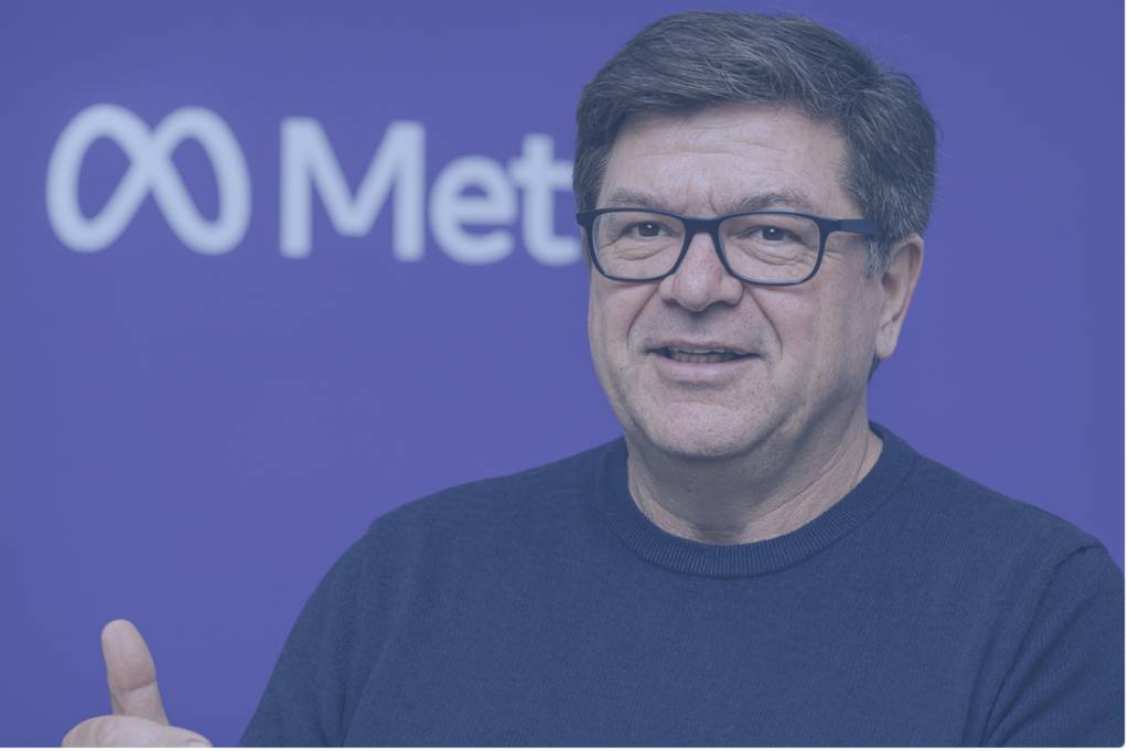 Portrait de Yann LeCun, directeur scientifique de l’IA chez Meta, souriant et s’exprimant devant un fond violet avec le logo Meta, vêtu d’un pull bleu marine et de lunettes noires.
