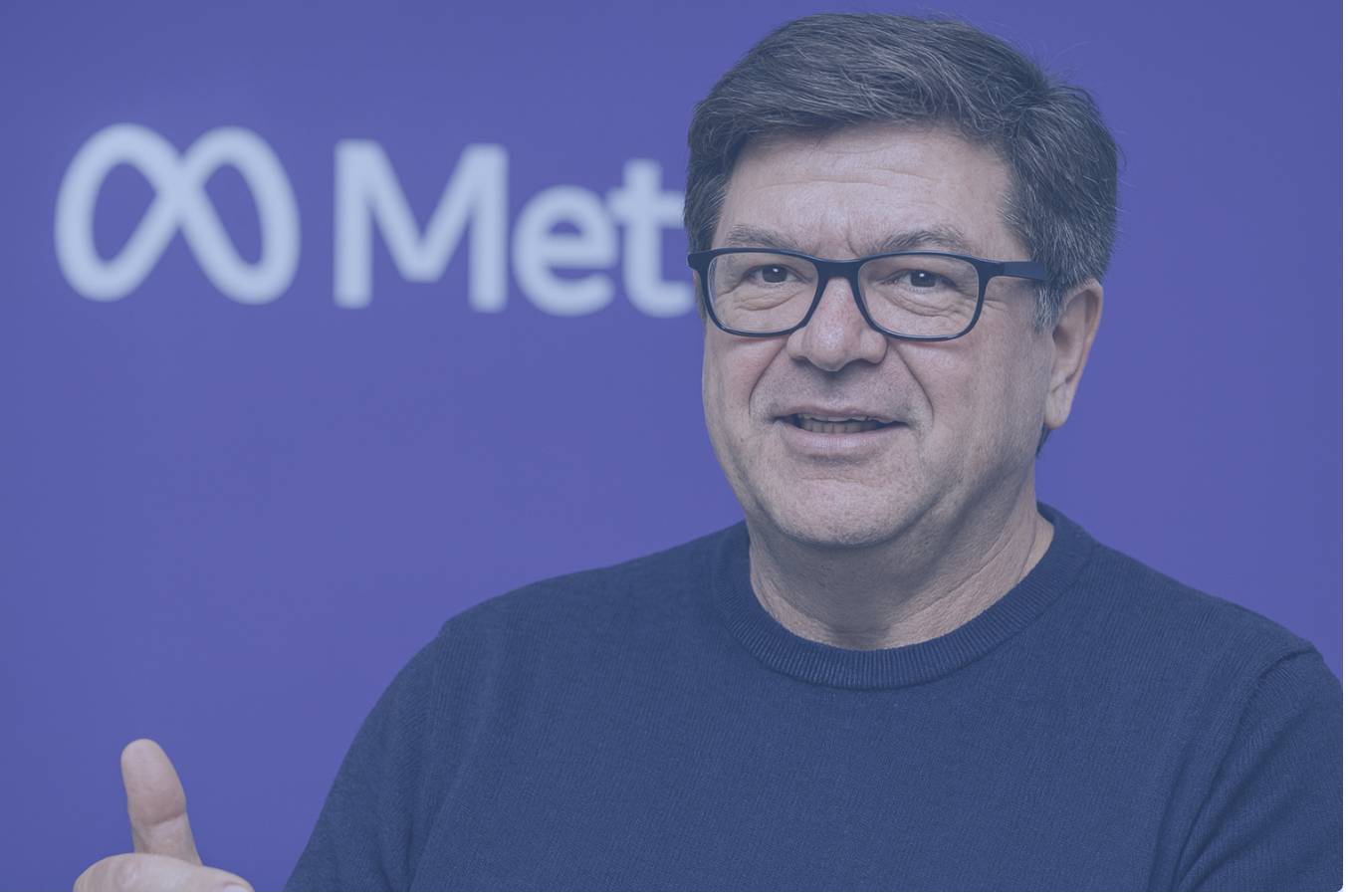 Portrait de Yann LeCun, directeur scientifique de l’IA chez Meta, souriant et s’exprimant devant un fond violet avec le logo Meta, vêtu d’un pull bleu marine et de lunettes noires.