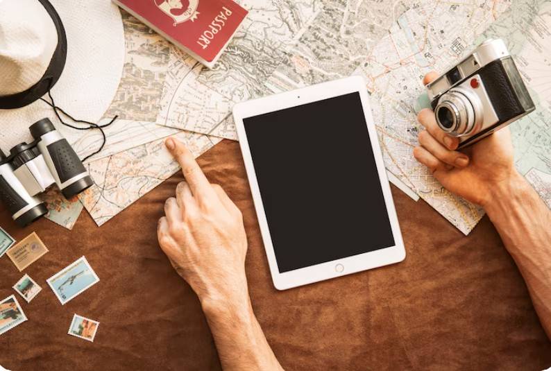 Vue de dessus d’une table avec une carte, une tablette, un passeport, un appareil photo, des jumelles et des timbres, représentant la préparation d’un voyage
