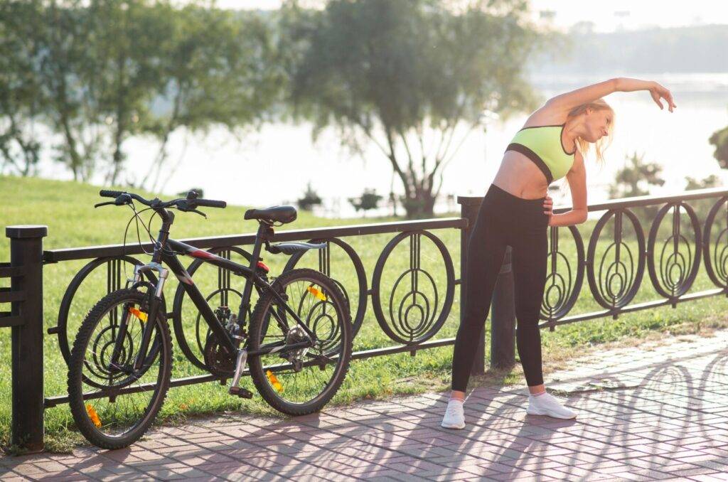Femme en tenue de sport s'étirant latéralement près de son vélo dans un parc ensoleillé, sur fond de rivière et d’arbres