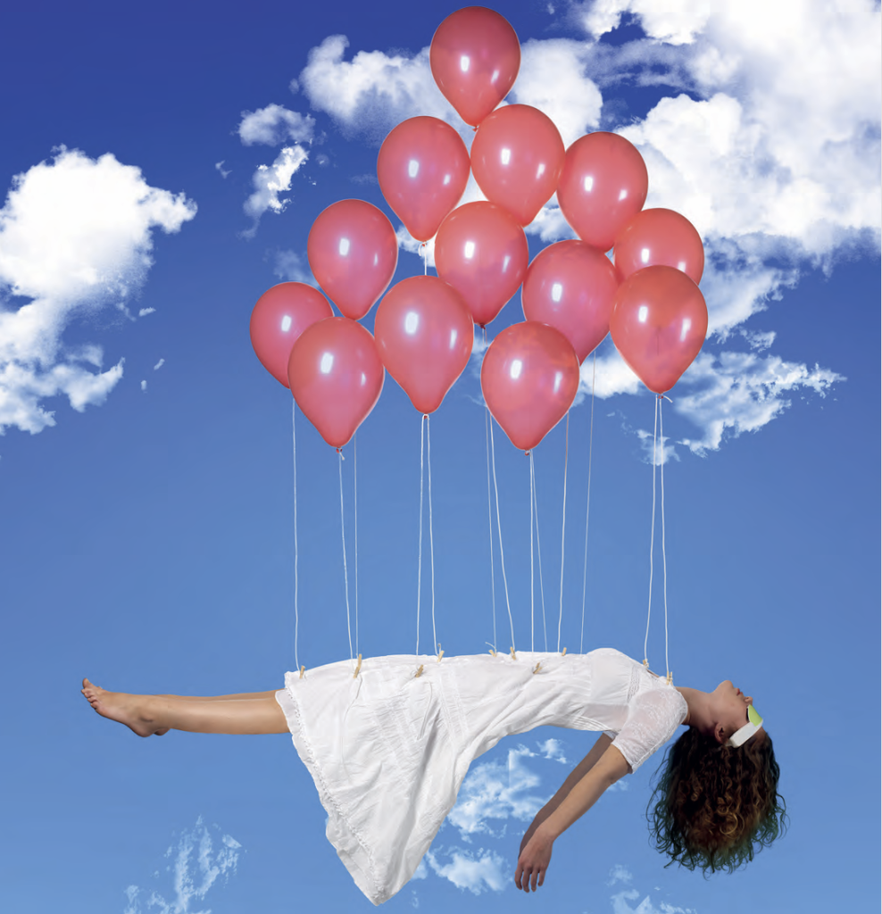 Femme allongée, les yeux fermés, flottant dans le ciel bleu grâce à des ballons roses attachés à sa robe, portant les lunettes de relaxation PSiO.