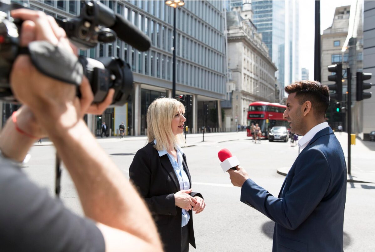 Interview d'une femme en costume par un journaliste avec micro et caméraman dans une rue passante avec un bus rouge en arrière-plan.